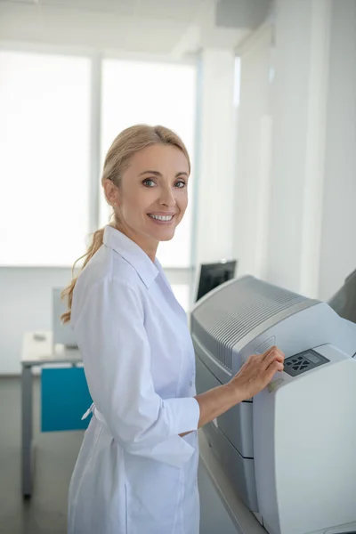 Blonde junge Ärztin steht im Büro und lächelt — Stockfoto