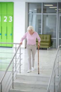 Elinde baston olan gri saçlı kadın aşağı iniyor.