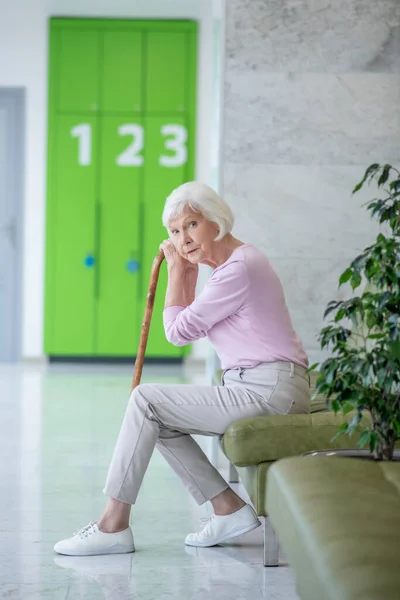 Grey-haired woman with a walking stick sitting in the corridor — Stock Photo, Image
