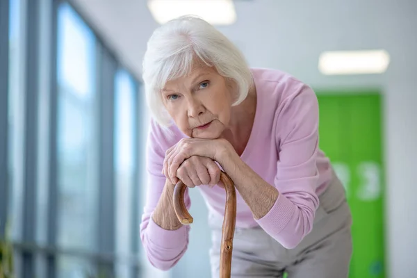 Grey-haired woman with a walking stick standing in the corridor and looking thoughtful — Stock Photo, Image