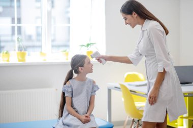 Koyu saçlı doktor, küçük hastasının ateşini ölçüyor.