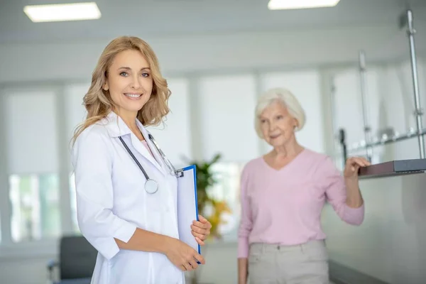 Sarışın gülümseyen doktor klinikte hastasının yanında duruyor. — Stok fotoğraf