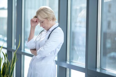 Yorgun kadın doktor, eli alnında pencerenin yanında duruyor.