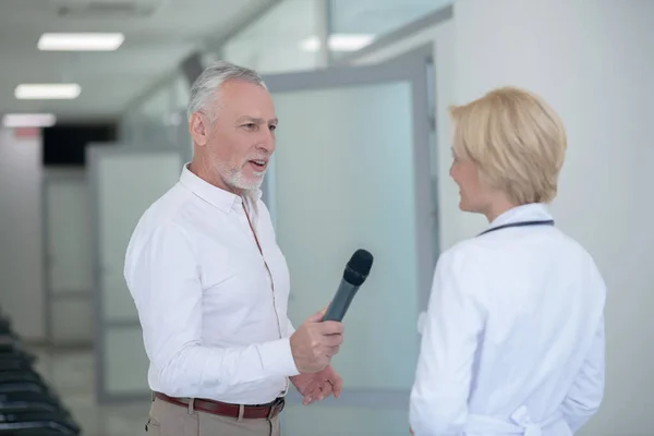 Periodista masculino de cabello gris entrevistando a doctora rubia en corredor — Foto de Stock