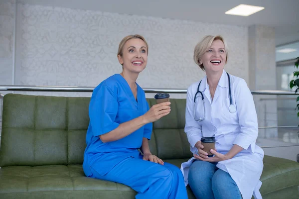 Дві жінки-лікарі сидять на дивані, насолоджуючись кавою — стокове фото
