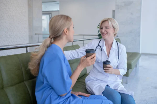 Dwie uśmiechnięte pracownice medyczne siedzące na kanapie, pijące kawę — Zdjęcie stockowe