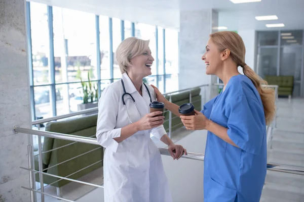 Дві усміхнені жінки-медики, спираючись на поручні сходів, п'ють каву — стокове фото