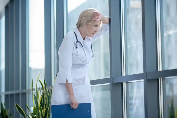 Втомлена жінка-лікар, що спирається на лоб на вікно — стокове фото