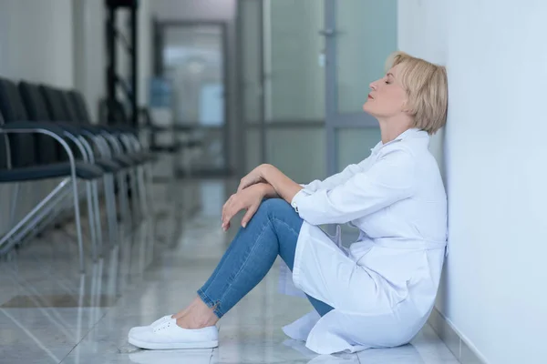 Cansada doctora rubia sentada en el suelo, apoyada en la pared — Foto de Stock