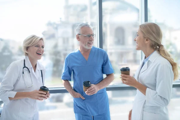 Group of smiling doctors drinking coffee next to the window, having friendly conversation — Stock Photo, Image