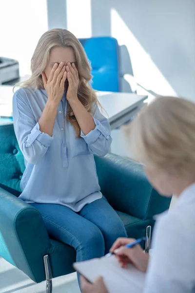 Aggódó női beteg eltakarja az arcát, szemben ül a női orvossal — Stock Fotó