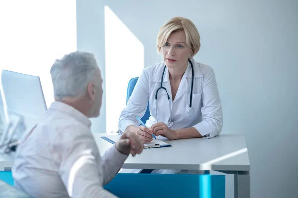 Sarışın kadın doktor masada oturuyor, yazı yazıyor, beyaz saçlı erkek hastaya danışıyor. — Stok fotoğraf