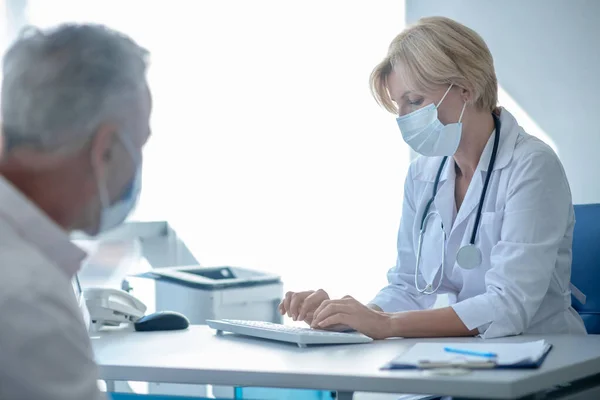 Жінка-лікар набирає маску, сіро-волохатий пацієнт сидить за столом навпроти неї — стокове фото