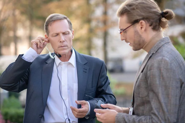 Volwassen man journawhile met headset passend oortje, zijn mannelijke collega helpen hem — Stockfoto