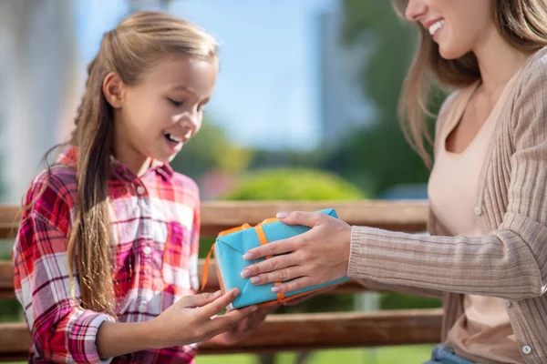 Мама дарит своей улыбающейся дочери коробку подарков на скамейке в парке — стоковое фото