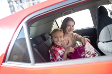 Gülümseyen kız ve annesi alışveriş torbalarıyla arka koltukta oturuyorlar.