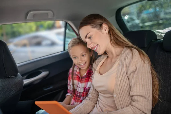 Memnun kız ve annesi arabanın arka koltuğundaki tabletten bir şeyi kontrol ediyorlar. — Stok fotoğraf
