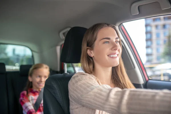 Sonriente mamá conduciendo con su hija sentada en el asiento trasero del coche — Foto de Stock