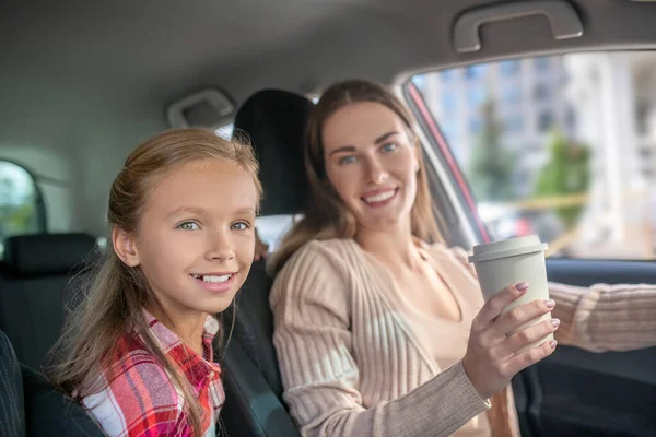 Gülümseyen anne elinde kahve fincanı, arabada kızıyla oturuyor. — Stok fotoğraf