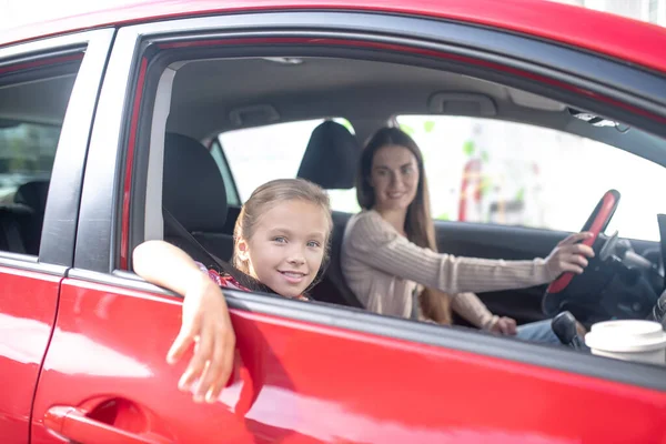 Smiling girl looking out the window on passenger seat, her mom sitting at the wheel — Stock Photo, Image