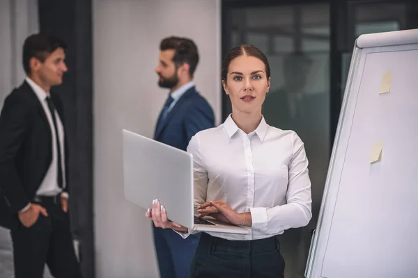 Mujer de cabello castaño de pie cerca del rotafolio con portátil, colegas masculinos hablando detrás — Foto de Stock