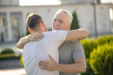 Genç kahverengi saçlı erkek ve olgun gri saçlı erkek birbirlerine sarılıyorlar.