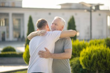 Kahverengi saçlı erkek ve olgun gri saçlı erkek birbirlerine sarılıyorlar, gülümsüyorlar.