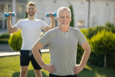 Gri saçlı erkek, elleri kalçasında, genç erkek, arkasında dambılları kaldırıyor.