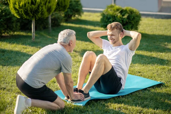 Gammal gråhårig hjälpa leende ung brunhårig hane med sit ups — Stockfoto