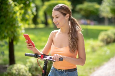 Turuncu bluzlu genç kız parkta scooter sürüyor ve bir mesaj okuyor.
