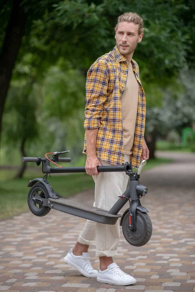 Man in checkered shirt holding his scooter and looking aside — Stock Photo, Image