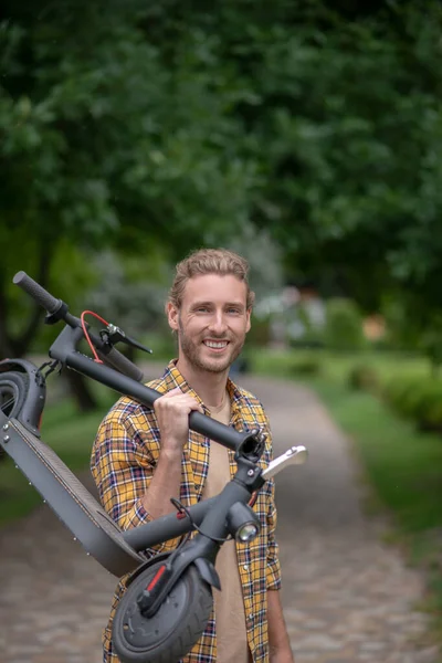 Homme en chemise à carreaux portant son scooter et souriant — Photo