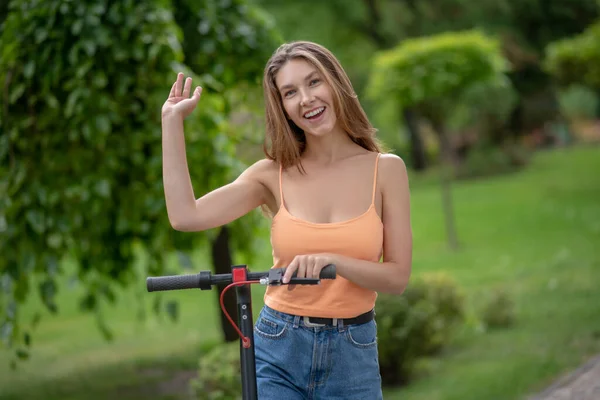 Cabelo comprido bonito menina montando scooter no parque e levantando a mão — Fotografia de Stock