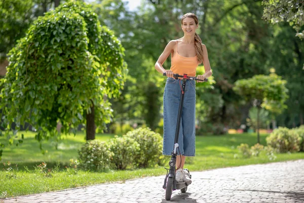Långhårig ung flicka rida en skoter i parken och ser nöjd — Stockfoto