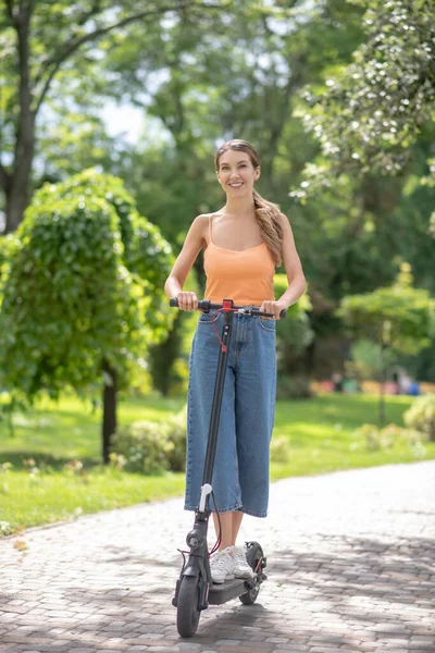 Giovane ragazza in top arancione cavalcando uno scooter nel parco e sorridendo — Foto Stock