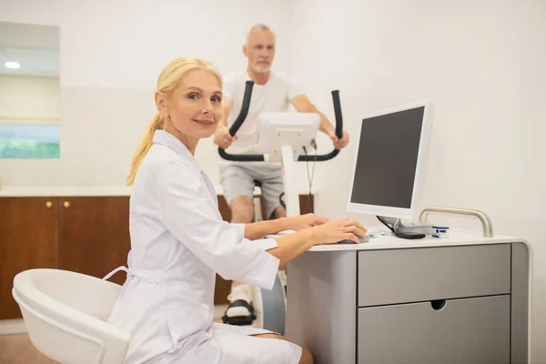 Médico loiro sentado no laptop enquanto seu paciente se exercita em uma esteira — Fotografia de Stock