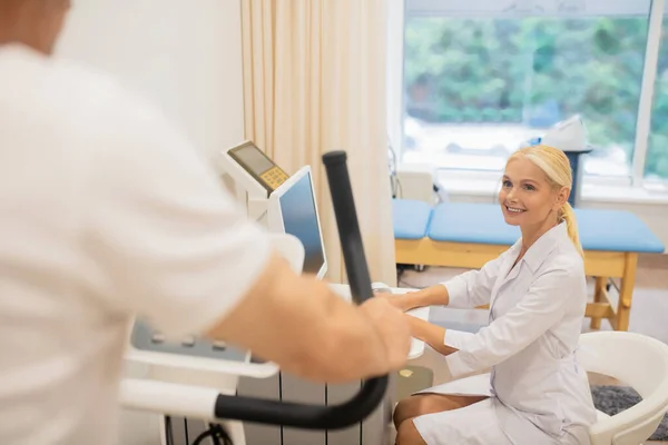 Ξανθιά χαμογελαστή γιατρός κάθεται στο φορητό υπολογιστή, ενώ ο ασθενής της άσκηση σε ένα διάδρομο — Φωτογραφία Αρχείου