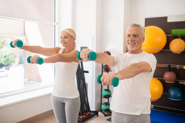 Hombre y mujer positivos haciendo ejercicio con mancuernas y extendiendo los brazos — Foto de Stock