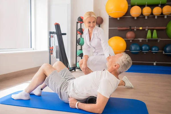 En kvinnlig sjukgymnast i labbrock som stöttar en patient och ler — Stockfoto