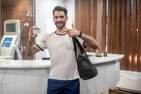 Bearded male standing at recepion desk, holding club card, pointing at it — Stock Photo, Image