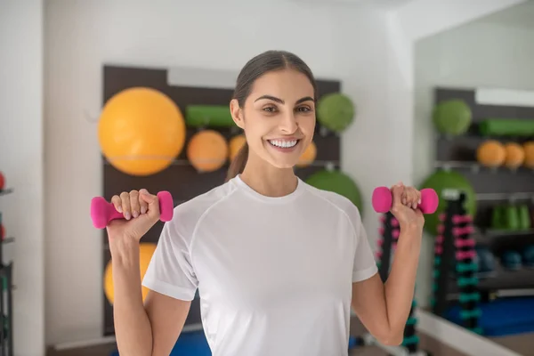 Sonriente hembra morena levantando pesas rosadas en el gimnasio — Foto de Stock