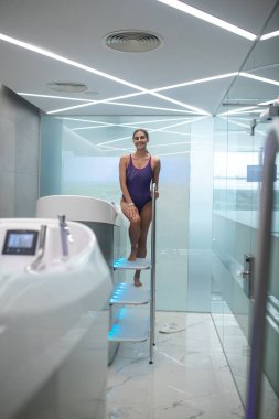 Gülümseyen siyah saçlı, mor mayo giymiş kadın hidromasaj küvetine giriyor.