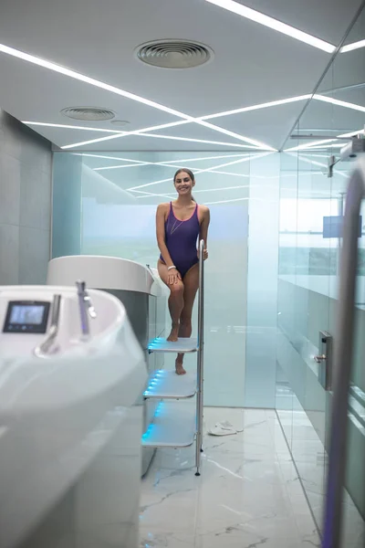 Smiling dark-haired female in purple swimming suit getting into hydromassage bathtub — Stock Photo, Image