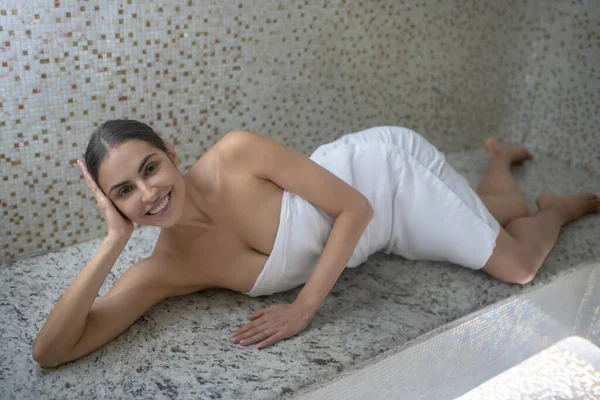 Donkerharig vrouwtje gewikkeld in witte handdoek liggend in sauna — Stockfoto
