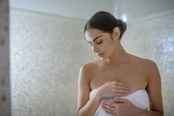 Dark-haired female standing in sauna, wrapped in towel, looking down — Stock Photo, Image