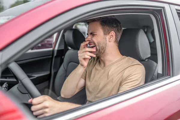 Уставший молодой мужчина за рулем красной машины, зевающий — стоковое фото