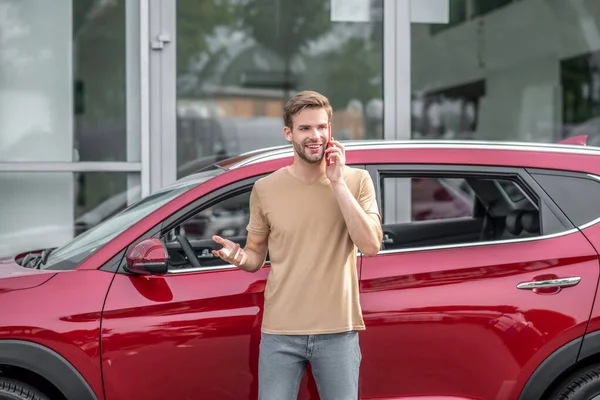 Mladý vousatý muž mluví na smartphonu před červeným autem — Stock fotografie