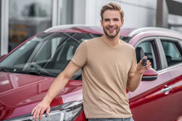 Sonriente joven macho apoyado en el coche rojo, sosteniendo las llaves — Foto de Stock