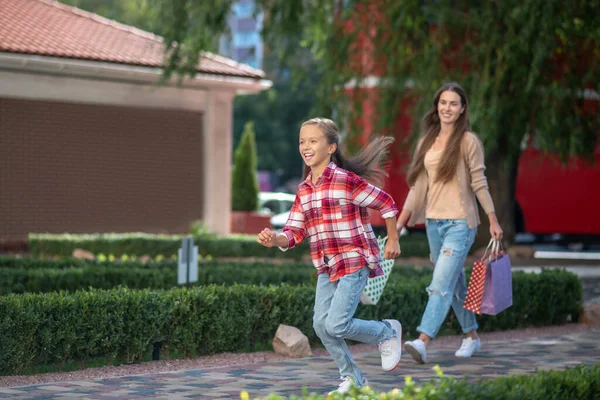 Adolescente hija corriendo delante de su mamá caminando con bolsas de compras — Foto de Stock