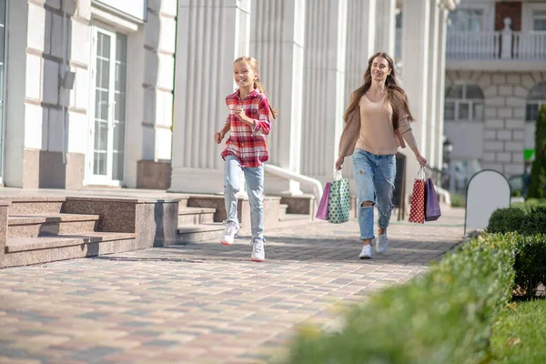 Szczęśliwa dziewczyna biegnie przed swoją mamą spacerując z torbami na zakupy — Zdjęcie stockowe